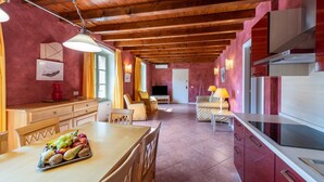 A spacious kitchen with wooden beams, a dining area with a fruit tray, and a living room with a TV and sofa. - Apartment 'Antica Torre D Annunzio' with Mountain View, Wi-Fi and Air Conditioning (San Felice del Benaco (BS))
