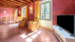 A room with a wooden ceiling, a television on a stand, a chair, a table, and a window with curtains. - Apartment 'Antica Torre D Annunzio' with Mountain View, Wi-Fi and Air Conditioning (San Felice del Benaco (BS))