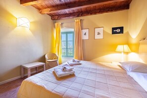 A hotel room with a bed, a chair, a small table, a window with curtains, and wall-mounted lights.