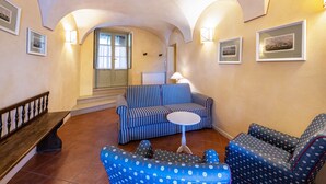 Una sala de estar con un sofá, sillones, una mesita y un banco. En las paredes hay cuadros enmarcados y una lámpara sobre la mesa.
