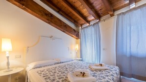 A bedroom with a wooden ceiling, a floral bedspread, two bedside tables with lamps, and a window with curtains. - Apartment 'Antica Torre Byron' with Shared Pool, Wi-Fi and Air Conditioning (San Felice del Benaco (BS))