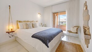A bedroom with a bed, bedside table, lamp, chair, and a view of the outside.