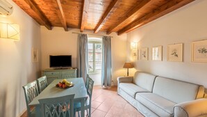 A room with a wooden ceiling, a dining table with chairs, a sofa, a TV, and framed pictures on the wall.