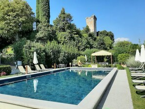 Une piscine rectangulaire bordée de chaises longues et d’ombrelles, entourée d’une végétation luxuriante et avec, en arrière-plan, une tour historique.