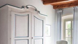 A room with a wooden ceiling, a white wardrobe with glass doors, a white armchair, and a bed with a floral bedspread.