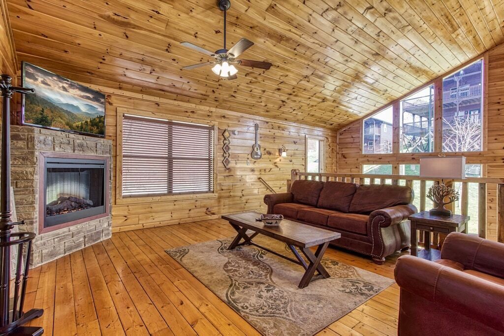 A log cabin interior with a fireplace, wooden floors, a brown leather sofa, and a ceiling fan.