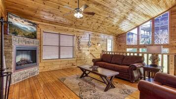 A log cabin interior with a fireplace, wooden floors, a brown leather sofa, and a ceiling fan.