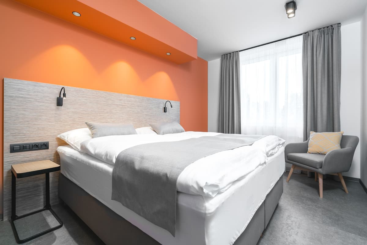 A modern hotel room with a large bed, a bedside table, a chair, and a window with curtains.