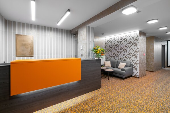 A reception area with a modern design, featuring a wooden reception desk, a grey sofa, and a patterned wall. - Hotel Diana (Karvina)