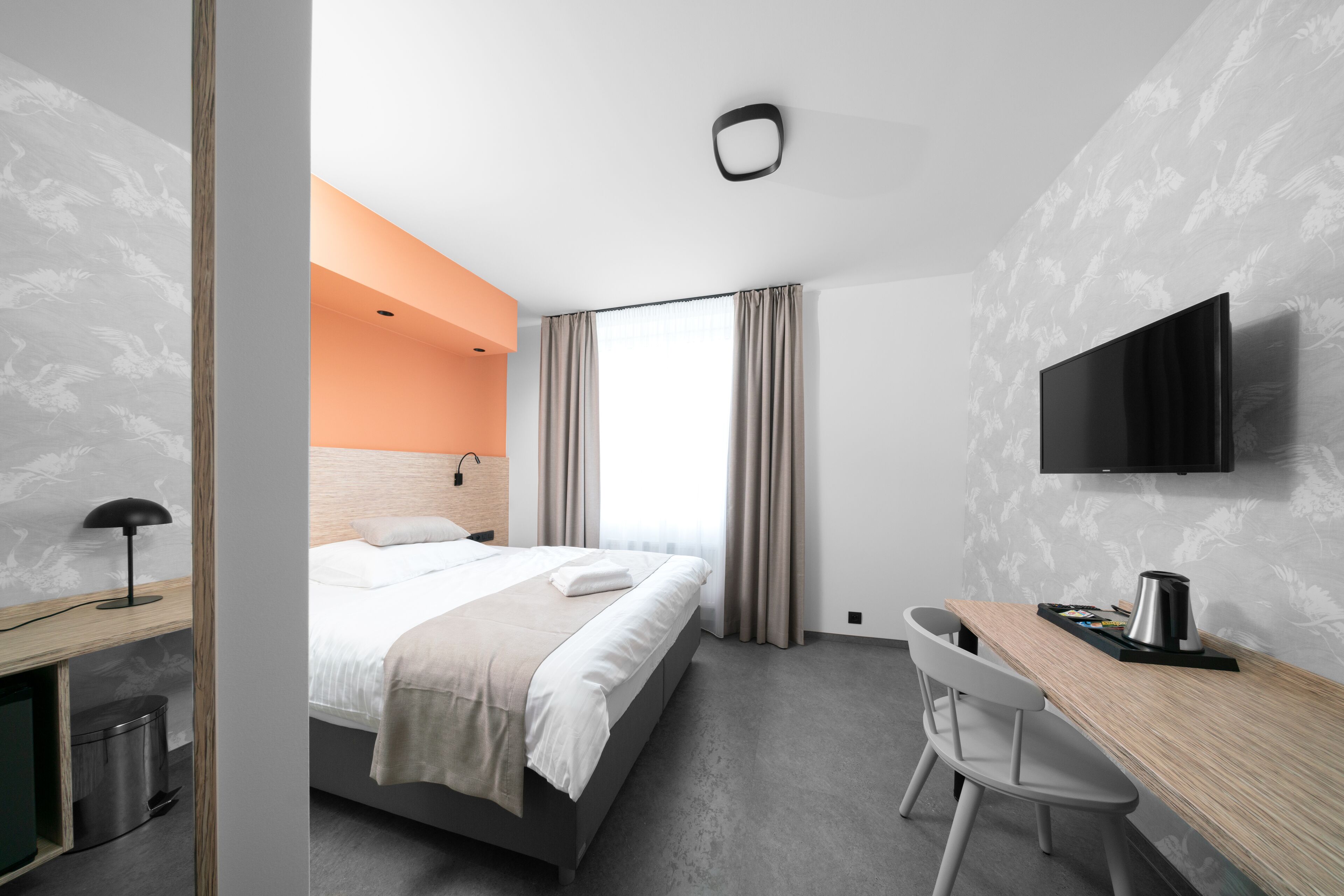 A modern hotel room with a bed, a desk, a chair, a TV, and a window with curtains.