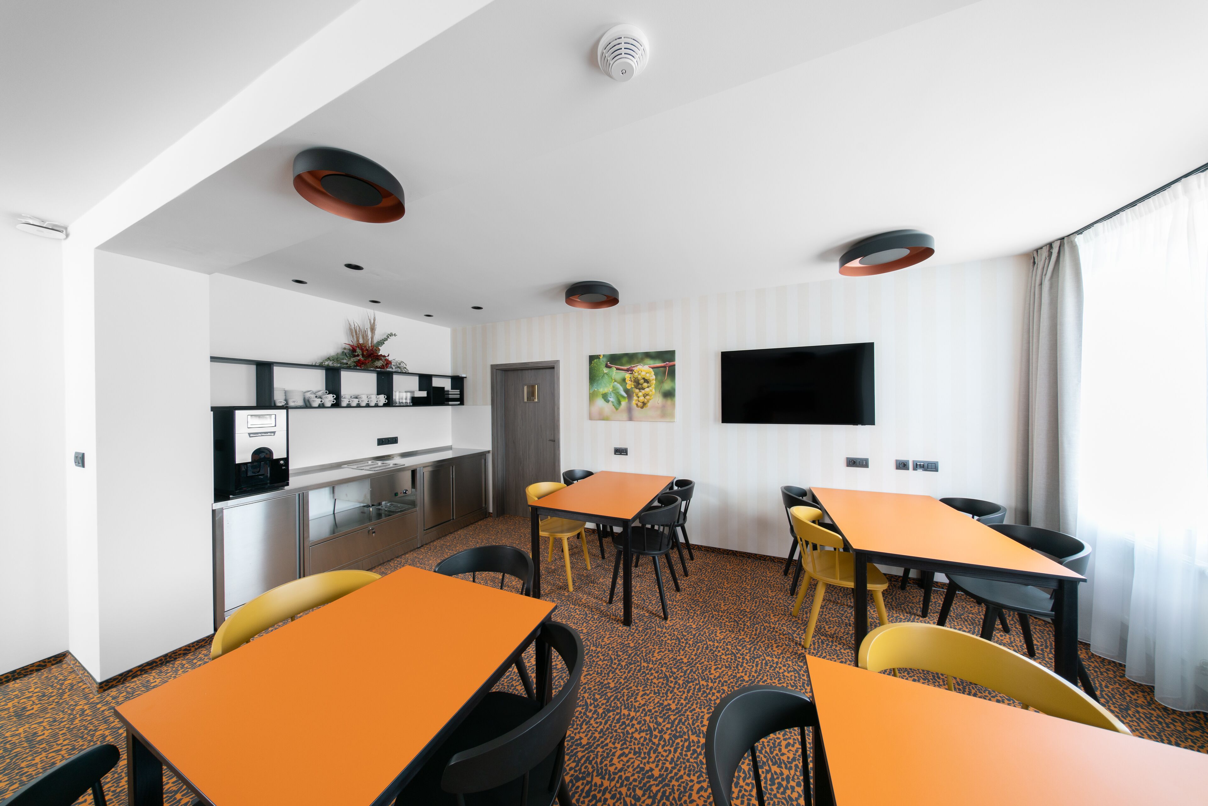 A modern hotel room with a kitchen area, a dining table with chairs, and a flat-screen TV mounted on the wall.