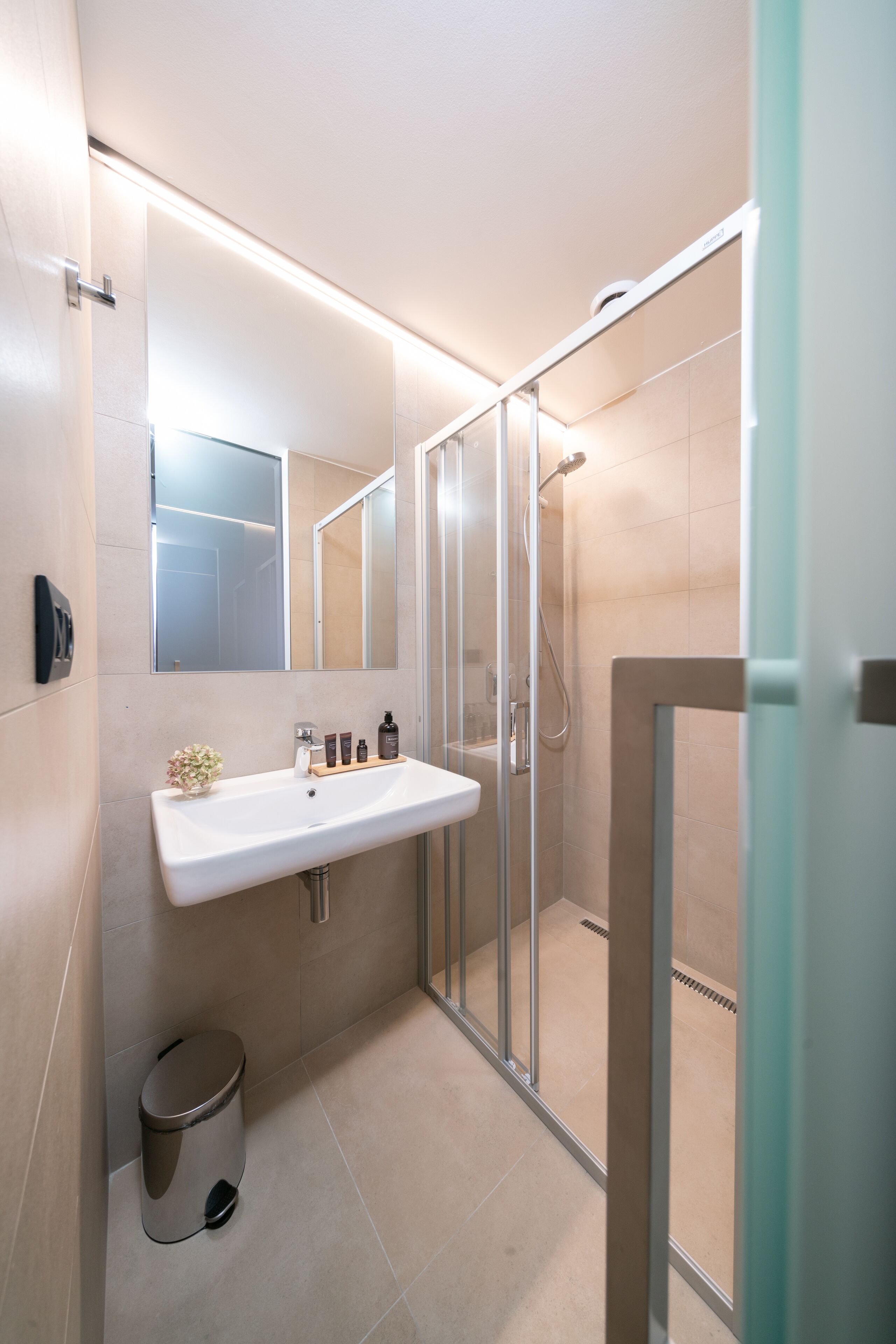 A modern bathroom with a glass shower enclosure, a white sink with a mirror above it, and a small trash bin.