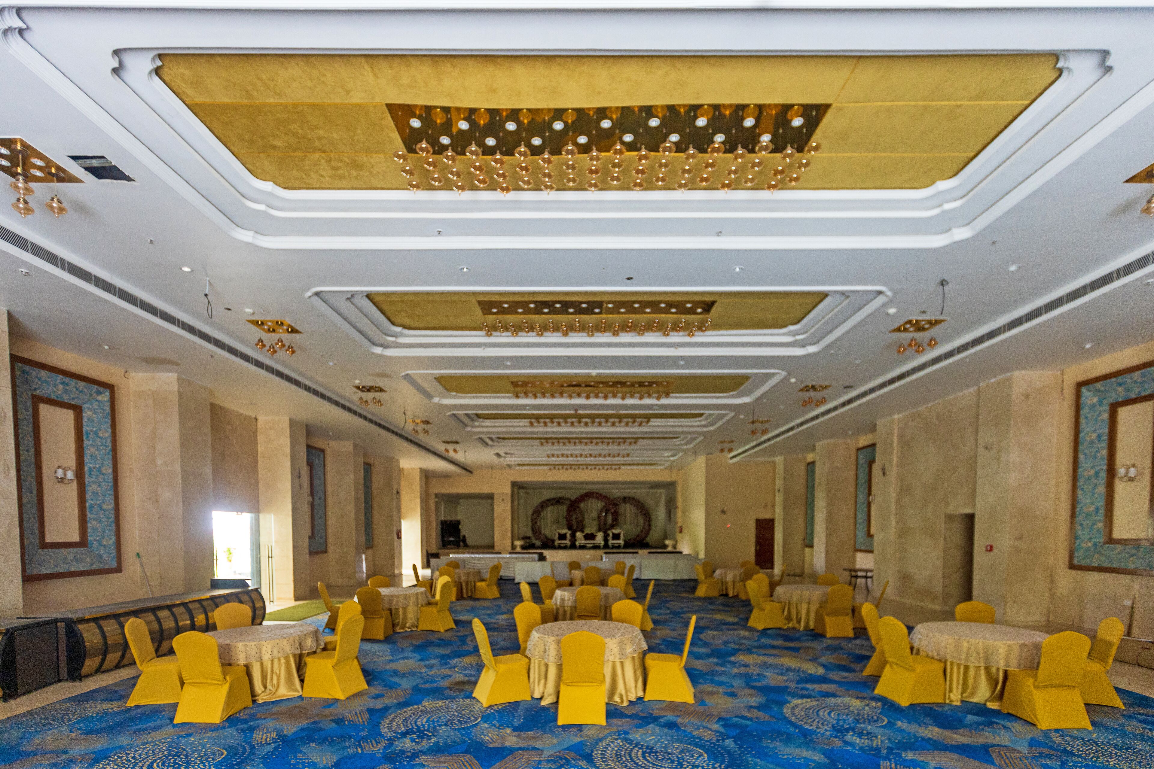 A banquet hall with round tables and yellow chairs, a blue carpet, and a decorative ceiling.