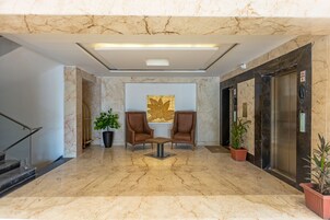 A lobby with marble flooring, two brown chairs, a table, potted plants, a staircase, and an elevator.