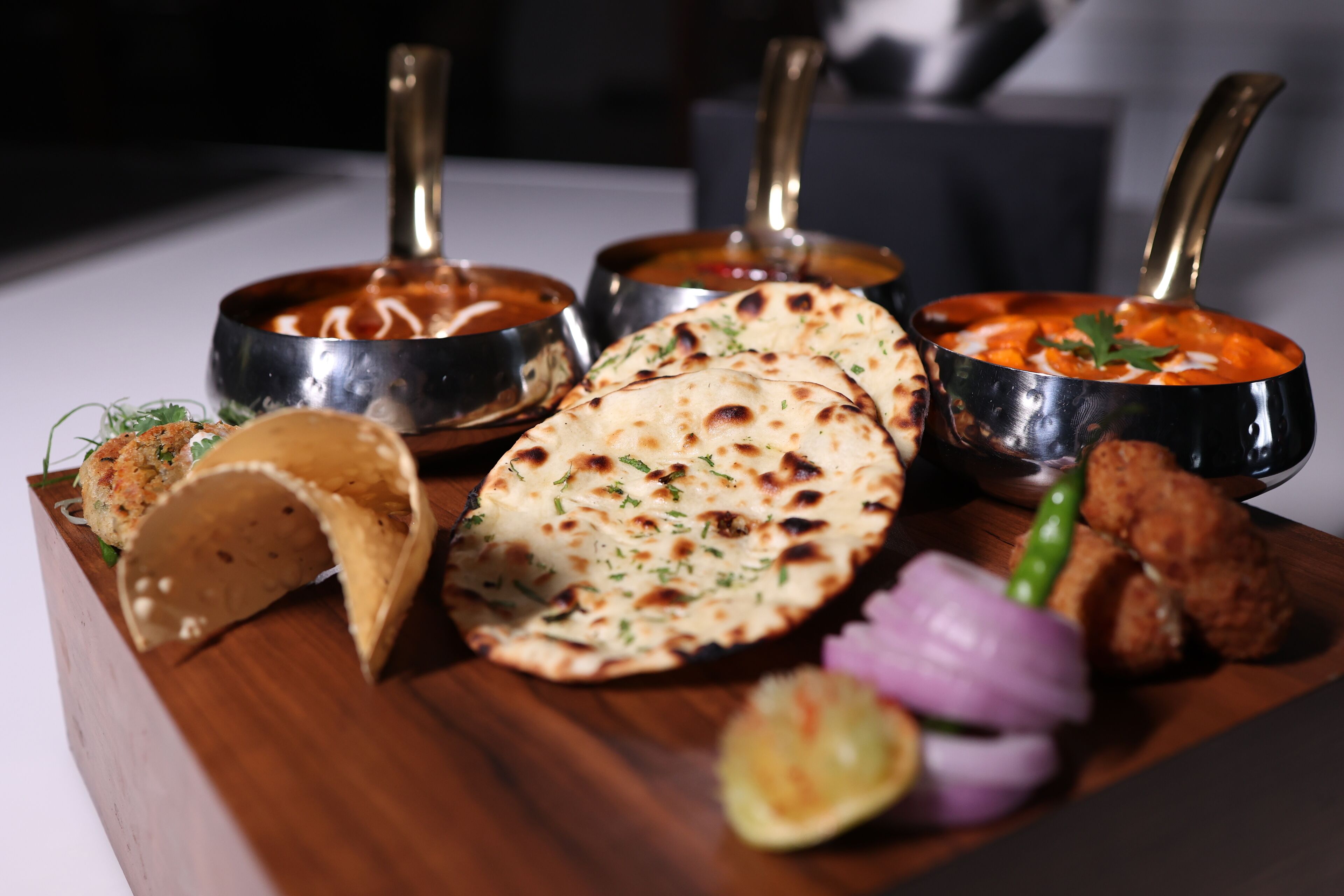 Un tagliere di legno con tre ciotole di curry in metallo, pane naan e un taco.