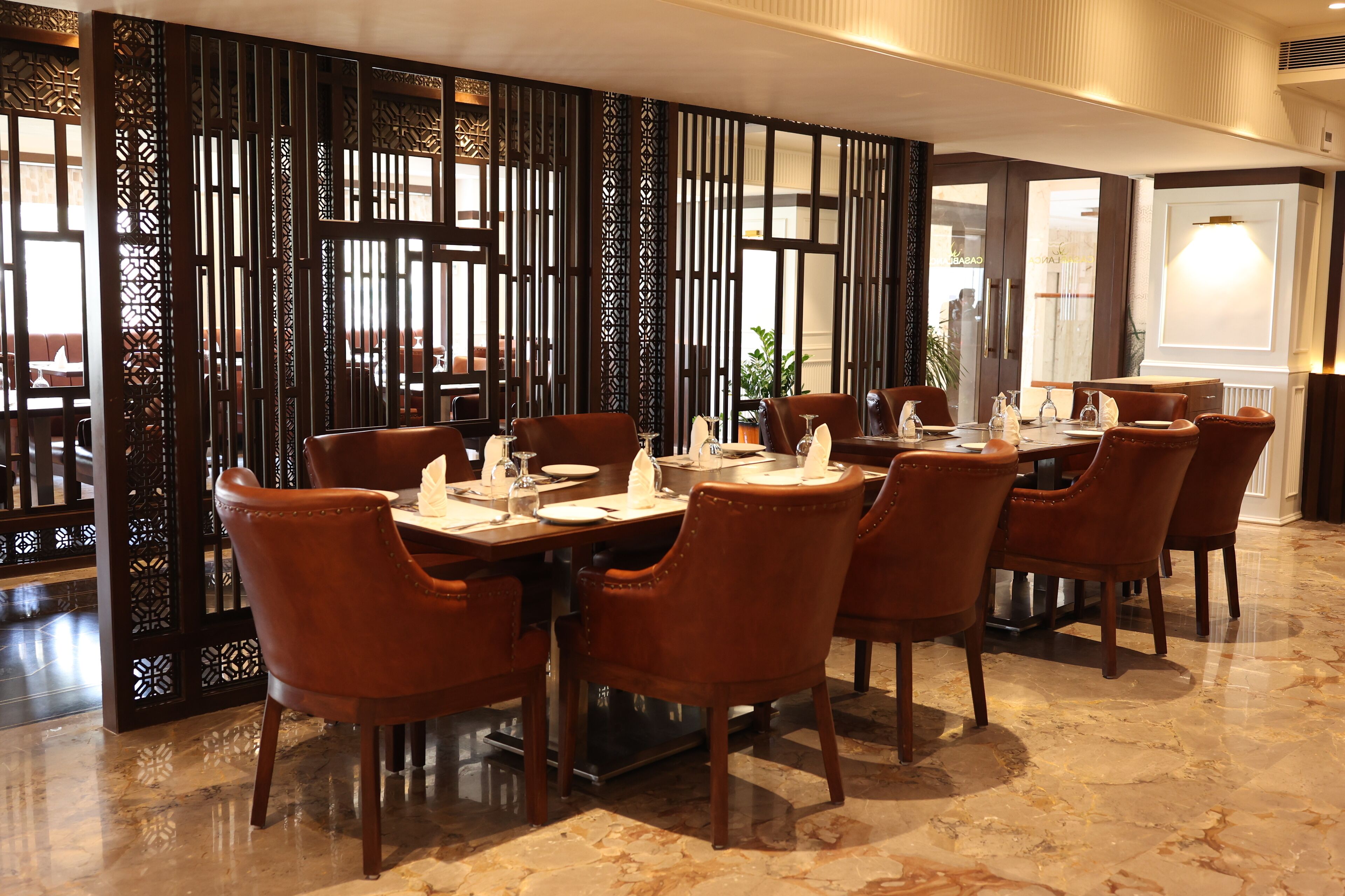 A dining area with a long table set for six, surrounded by six brown leather chairs. The floor is tiled with a marble pattern.