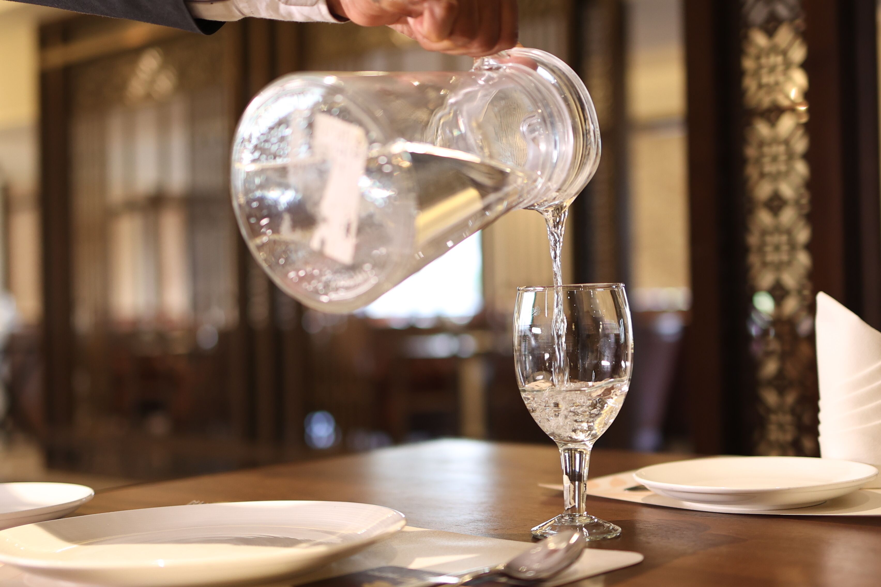 A person pouring water from a pitcher into a glass on a table.