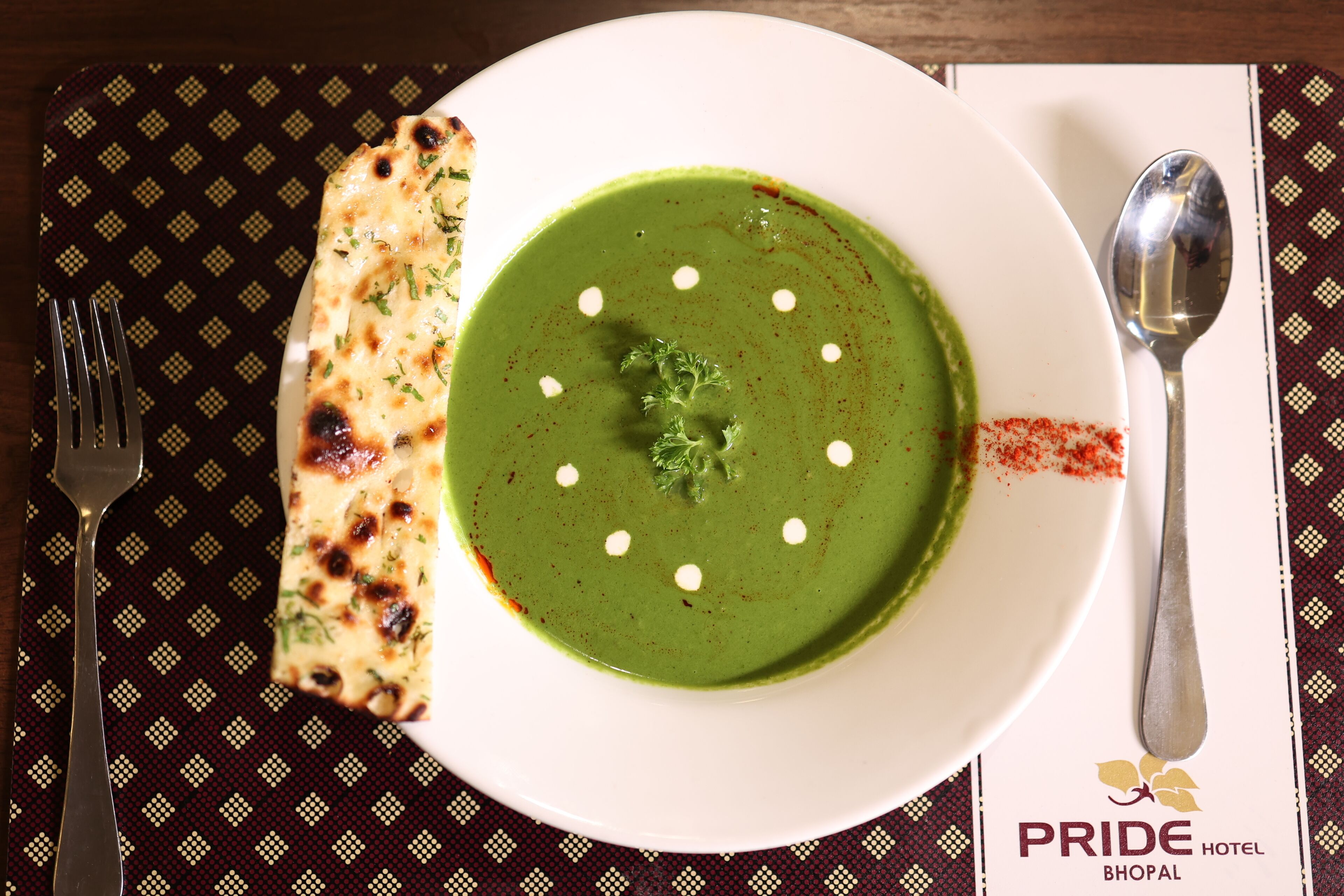 Une assiette de soupe verte, garnie de cuillerées de crème et accompagnée d’un morceau de pain naan, le tout servi sur une assiette blanche avec une cuillère et une fourchette, posé sur un set de table à motifs.