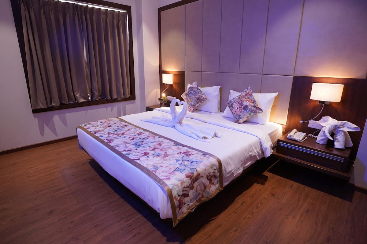 Una habitación de hotel con una cama grande, suelo de madera, mesita de noche con lámpara y ventana con cortinas.