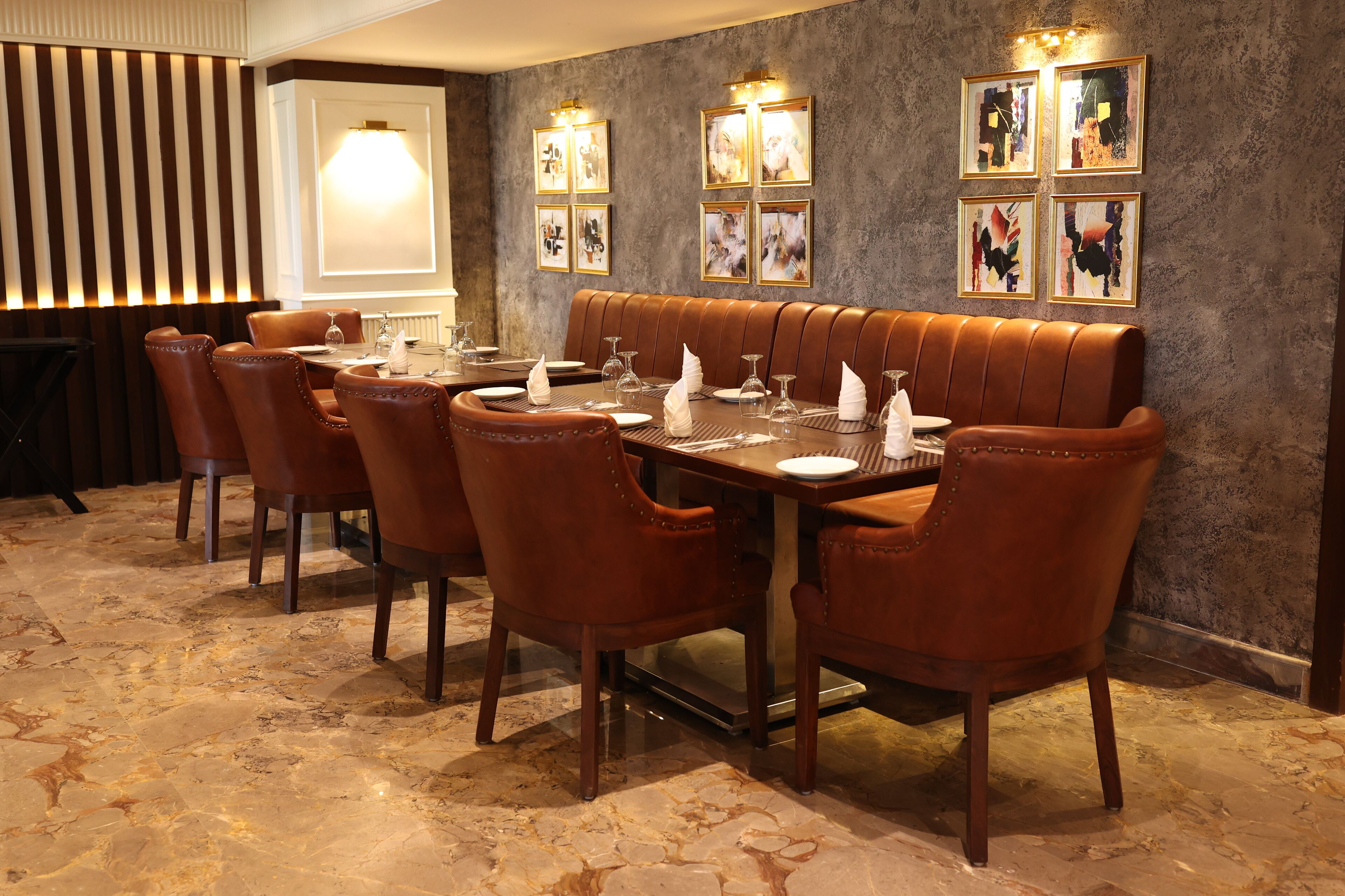 A dining area with a long brown leather booth and wooden chairs, marble flooring, and framed artwork on the walls.