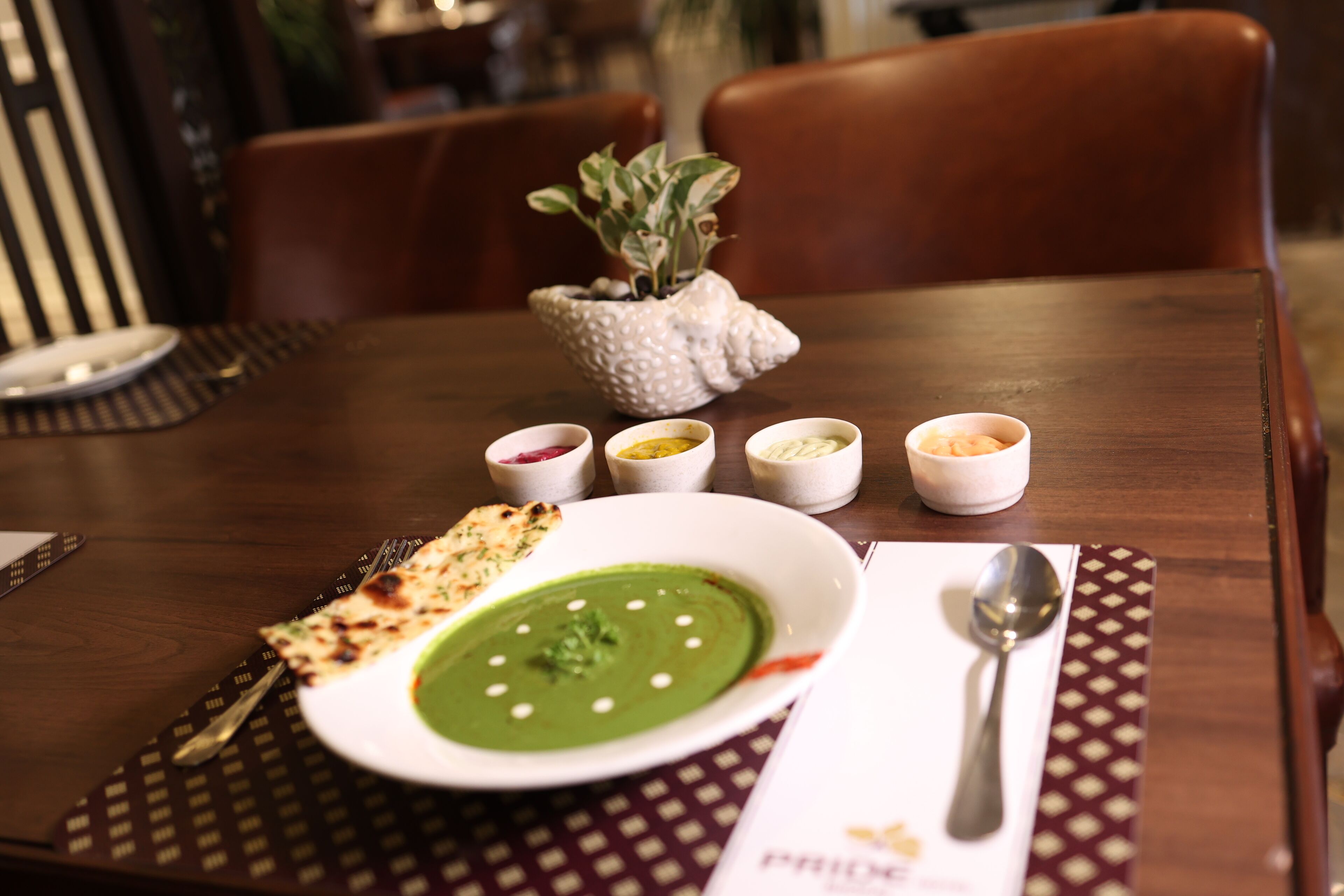 A dining table with a plate of green soup, several small bowls of condiments, a spoon, and a piece of naan bread.