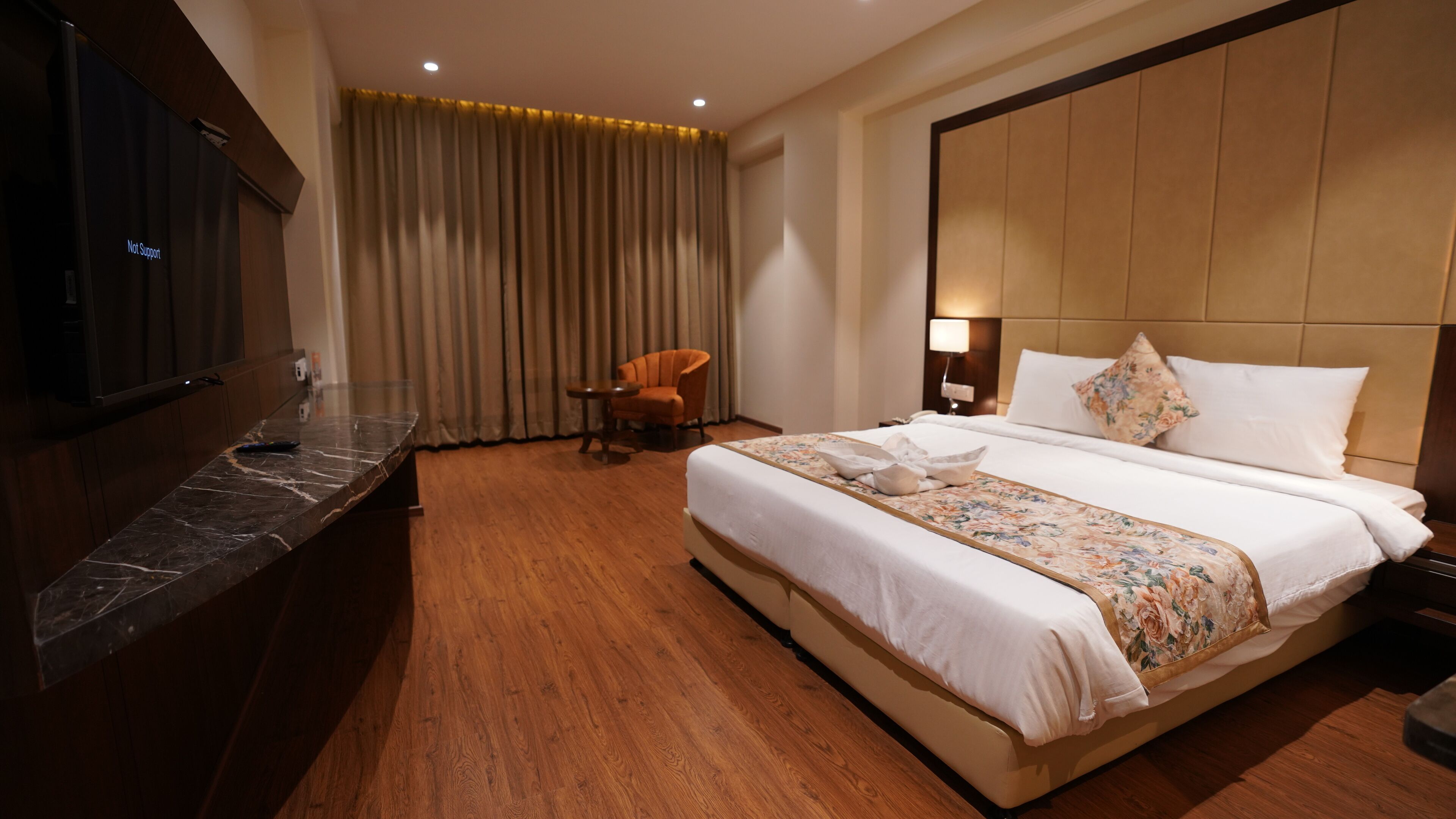 Una habitación de hotel con una cama grande, piso de madera, televisor a la izquierda y una silla cerca de la ventana.