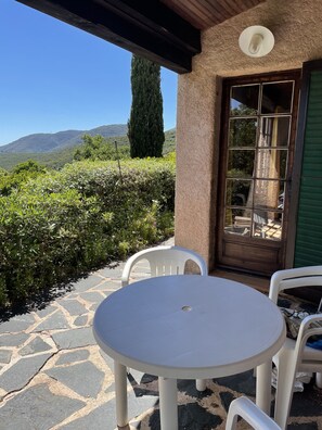 A patio with a round table and two chairs, a door with a window, and a view of a mountain and greenery. - Vacation home with a beautiful ocean view (La Croix-Valmer)