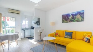 Ein hellgelbes Sofa mit gemusterten Kissen, ein Esstisch mit Stühlen, ein kleiner weißer Beistelltisch und ein Küchenbereich mit Herd und Schränken.