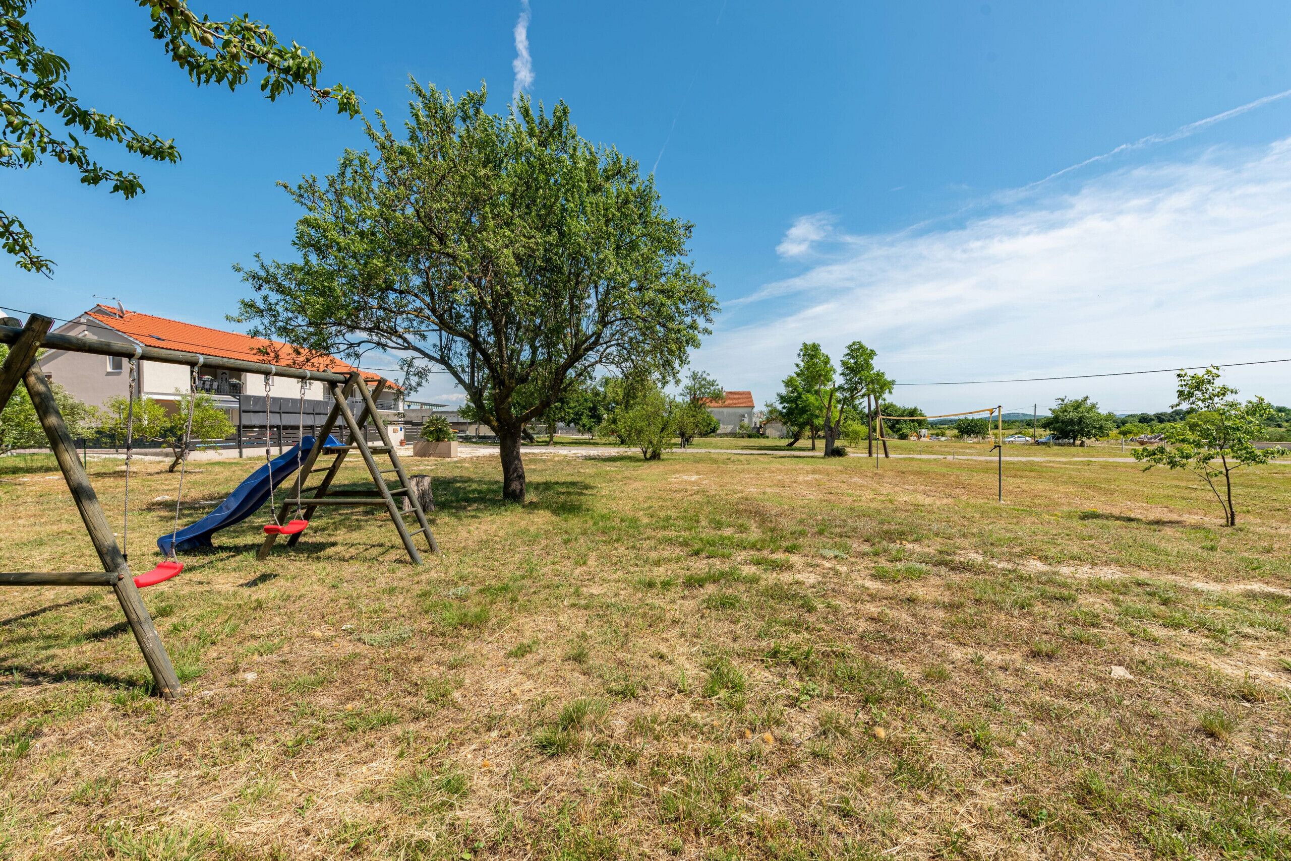 A playground with a slide and swings, surrounded by grass and trees, with houses in the background.