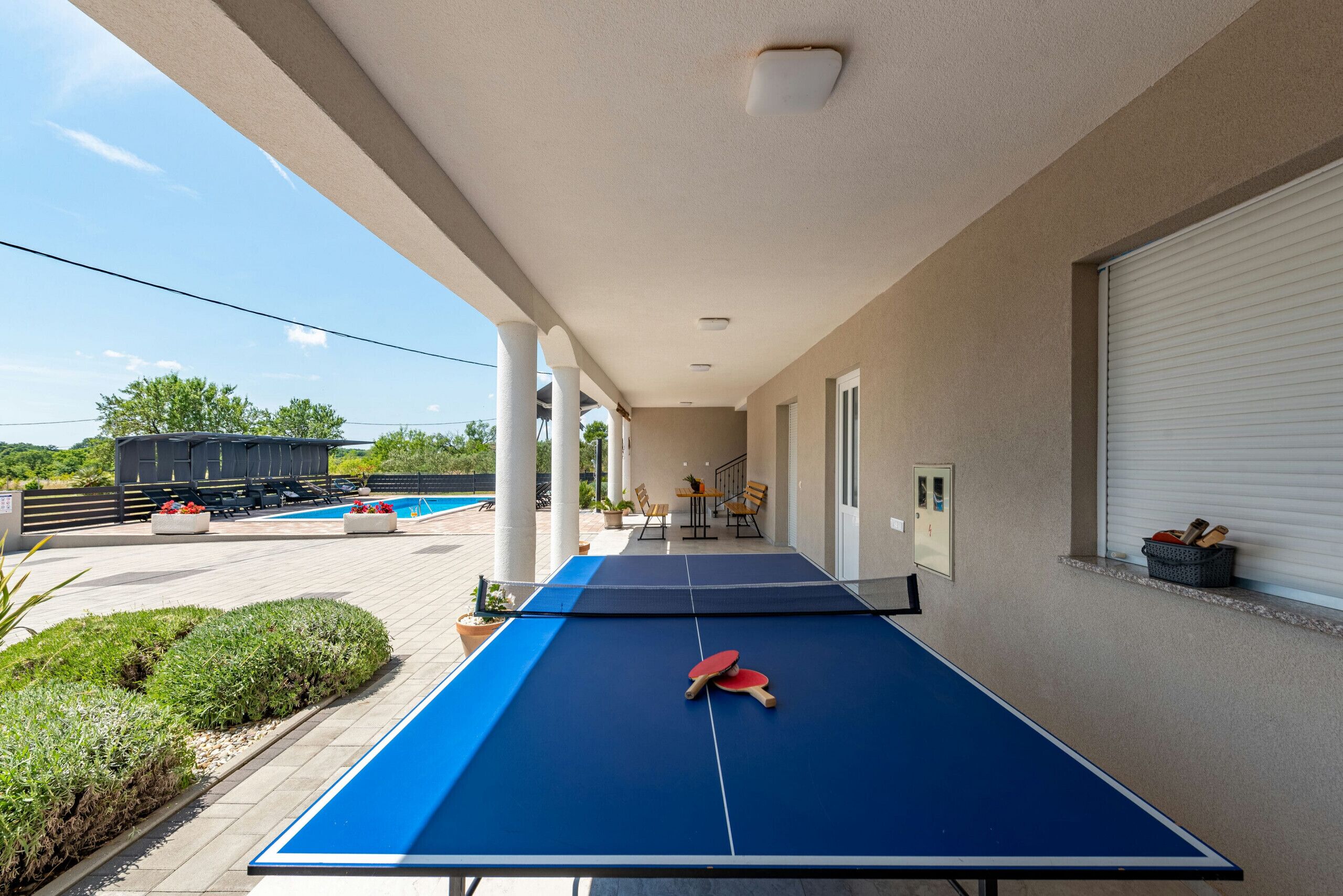 An outdoor ping pong table with two red paddles, surrounded by a pool area with lounge chairs and a staircase leading to an upper level.