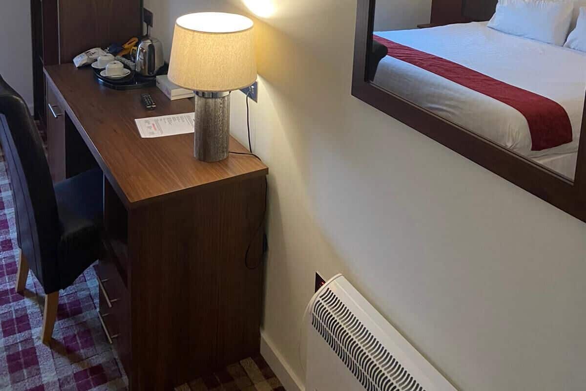 Habitación de hotel con cama, escritorio con lámpara, silla, armario, televisor de pantalla plana y radiador.