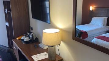 Quarto de hotel com escrivaninha, luminária, televisão, cama e espelho refletindo o ambiente.