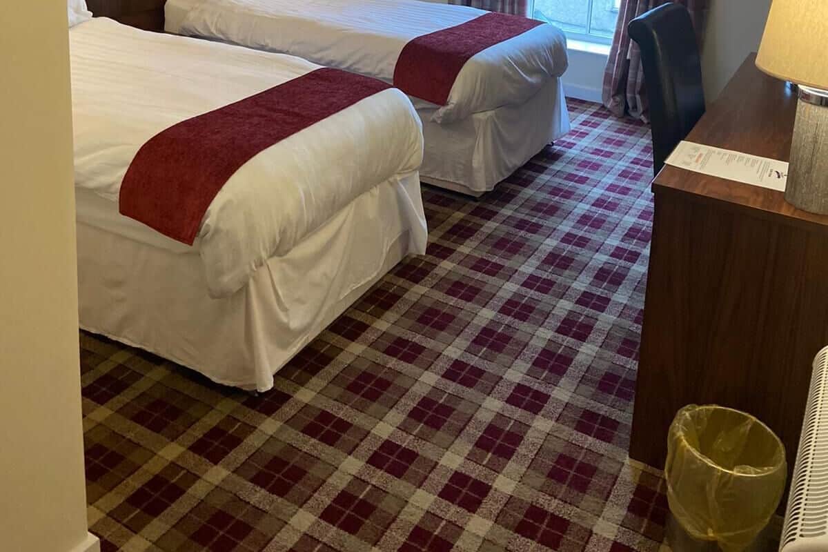 Habitación de hotel con dos camas, piso a cuadros, ventana con cortinas, un escritorio con lámpara y un cubo de basura.