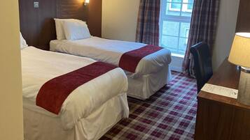 A hotel room with two beds, a checkered floor, a window with curtains, a desk with a lamp, and a trash bin.