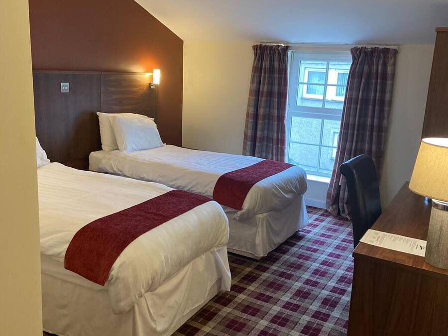 A hotel room with two beds, a checkered floor, a window with curtains, a desk with a lamp, and a trash bin.