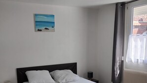 Una cama bien tendida en una habitación con un cuadro de playa en la pared, una ventana con cortinas y un radiador.