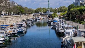 A canal with boats docked along the sides, a bridge in the background, and a statue on a tall pedestal.