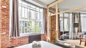 A hotel room with a large bed, a window with curtains, a brick wall, and a sliding glass door leading to a balcony. - NEW Design Flat in the Heart of Paris-An Ecoloflat (Paris)