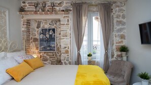 Ein Schlafzimmer mit Steinwand, einem Bett mit gelb-weißer Bettwäsche, einem grauen Sessel und einem Bild an der Wand.