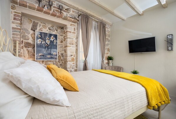 A bedroom with a stone wall, a painting, a bed with white and yellow bedding, a TV, and a clock.