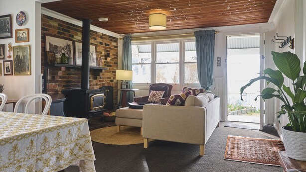 A cozy living room with a fireplace, a sofa, a dining table, and a large window with curtains. - gold miners cottage (Lancefield)