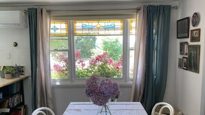 A dining area with a table set for two, a vase with flowers, a bookshelf, and a window with curtains. - gold miners cottage (Lancefield)