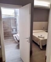 Una habitación de hotel moderna con suelo de madera, una cama con edredón blanco y un baño con cabina de ducha de vidrio, con vistas al exterior.