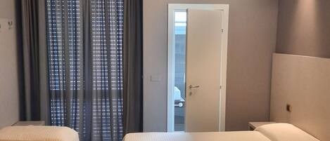 A hotel room with two beds, a window with curtains, a door, and a view of the outside.