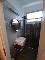 A modern bathroom with a rectangular sink, a round mirror, a glass-enclosed shower, and a window providing natural light.