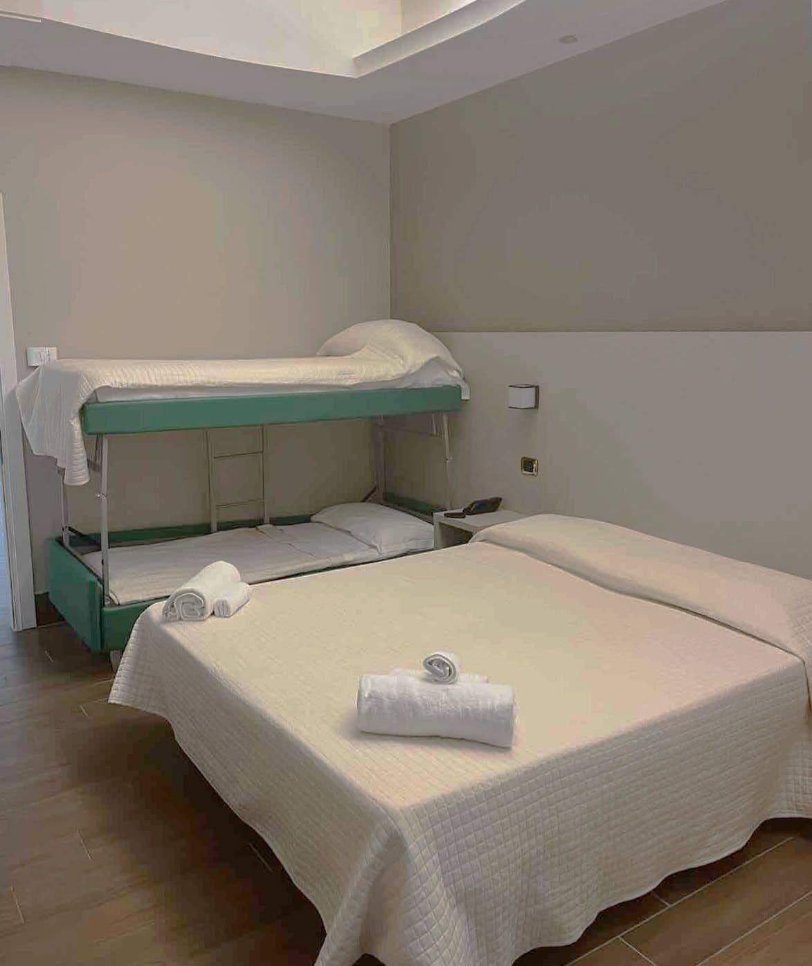 Habitación con literas de estructura verde y ropa de cama blanca. Se ve una mesita de noche pequeña con una lámpara y una toalla doblada sobre la cama.