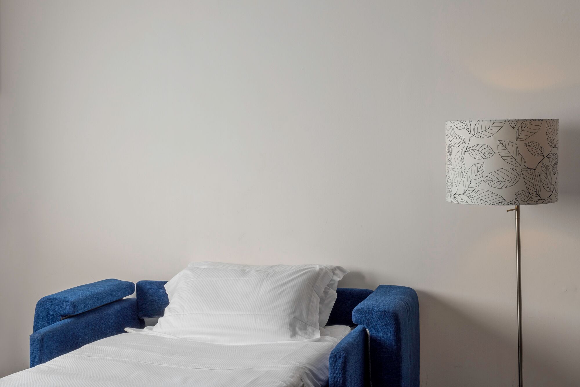 A bed with white bedding and a blue headboard, a floor lamp with a leaf pattern, and a plain wall.