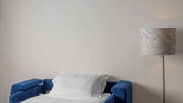 A bed with white bedding and a blue headboard, a floor lamp with a leaf pattern, and a plain wall.