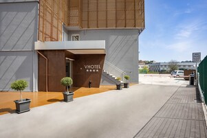 Una entrada de hotel moderna con una escalera que sube al segundo piso, plantas en macetas y cielo despejado.