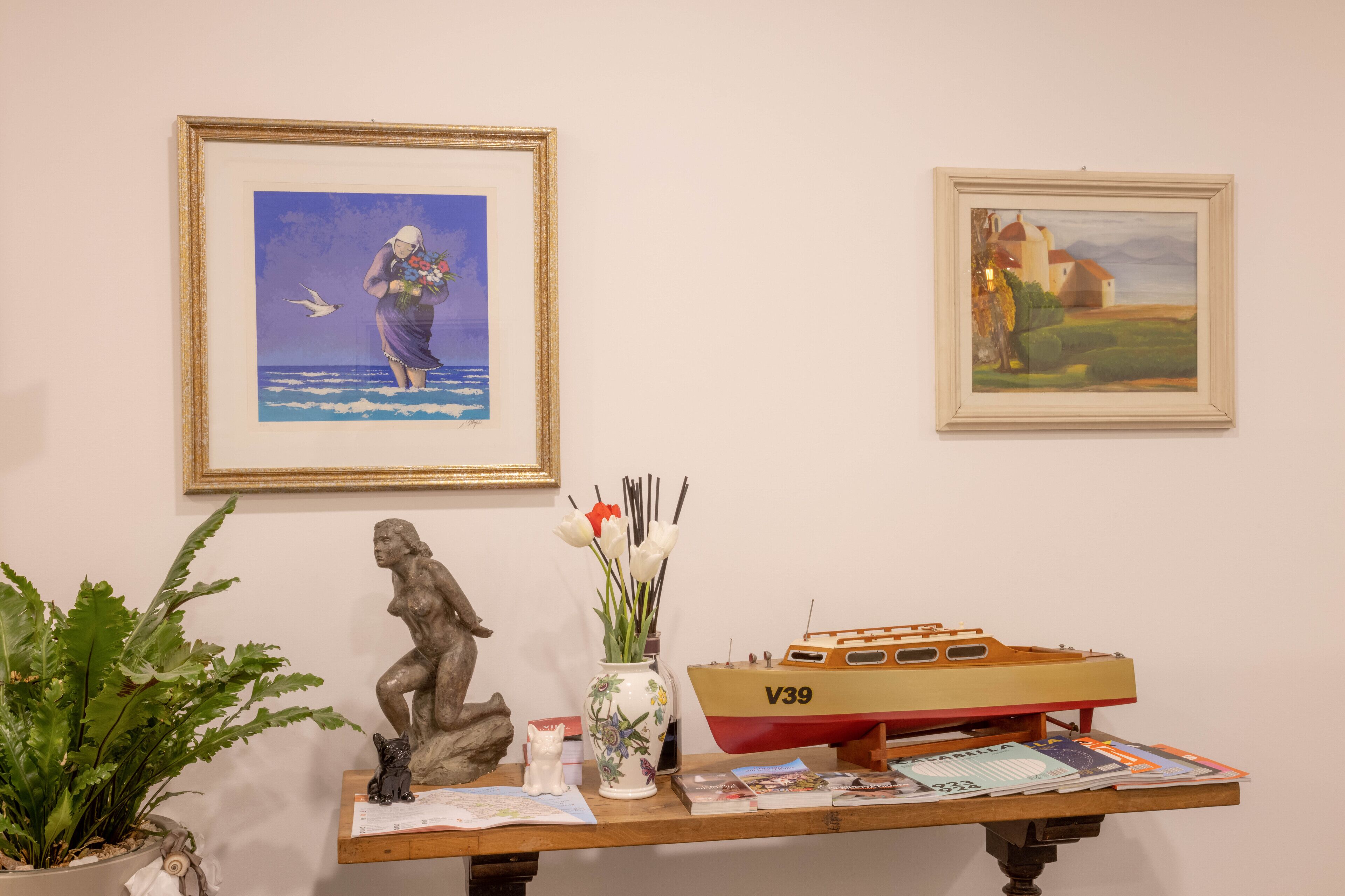 Un tavolo con un quadro incorniciato, un vaso con fiori, un modellino di barca e una pianta in vaso.